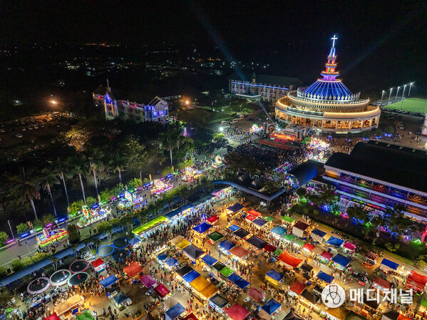 Aerial view of the Sakon Nakhon Christmas Star Parade showing illuminated star processions, festive market stalls, and the Cathedral of St Michael the Archangel during the annual celebration in Tha Rae, Sakon Nakhon.